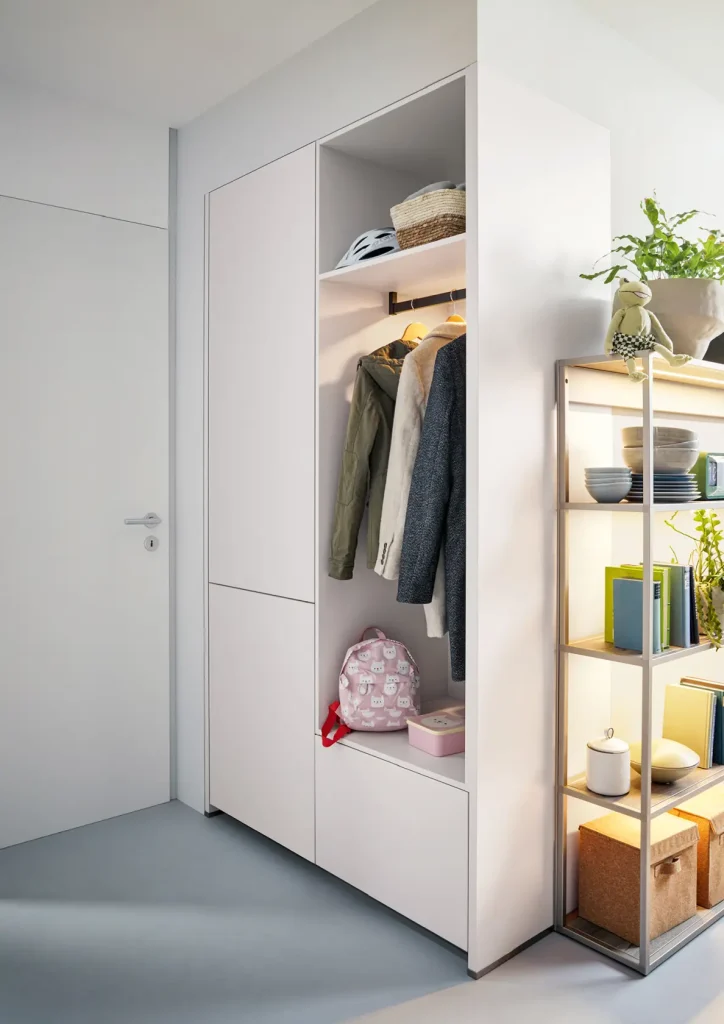 Ein moderner Flurschrank mit Mänteln, einem rosa Rucksack, Körben und Hüten steht neben eleganten Wohnmöbeln. Daneben stehen auf einem hellen Holzregal Pflanzen, Bücher und Deko-Schachteln. Das Bild wirkt aufgeräumt und gut organisiert.