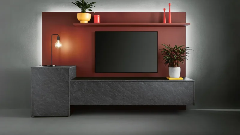 Moderner TV-Schrank mit dunkler Steinoptik, einem an der Wand montierten Flachbildfernseher, einer roten Rückwand, einem Regal mit Pflanzen und roten Dekoelementen sowie einer Seitenlampe auf einem grauen Schrank. Die minimalistische und hell beleuchtete Einrichtung besticht durch stilvolle Wohnmöbel-Elemente, die Ihrem Wohnraum sowohl Funktionalität als auch Eleganz verleihen.