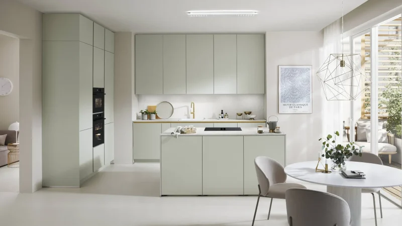 Moderne Küche mit hellgrünen Schränken, einer Kochinsel mit Kochfeld, Einbaubacköfen und einem Essbereich mit einem runden Tisch und vier Stühlen. Ein großes Fenster lässt Tageslicht herein und schafft eine einladende Wohnatmosphäre. Die minimalistische Einrichtung und die durchgehend neutralen Farbtöne unterstreichen das Gefühl von Komfort und Stil.