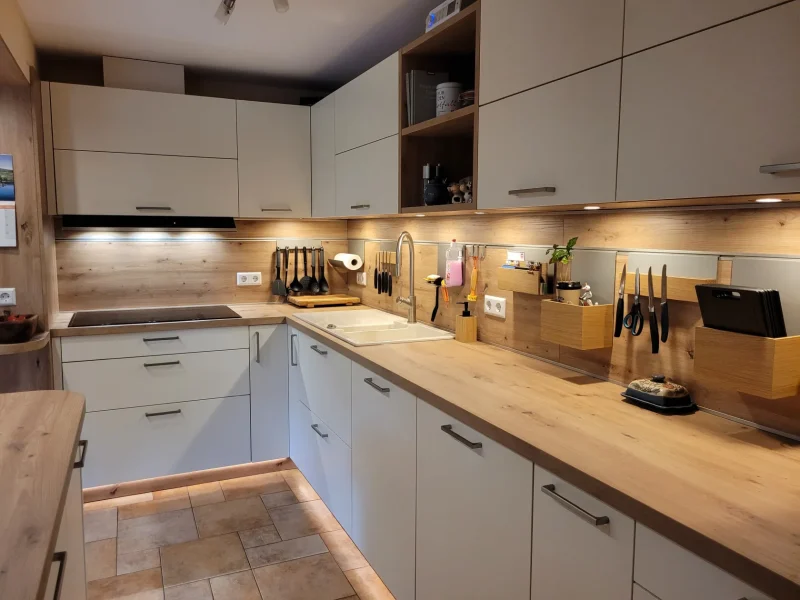 Eine moderne Küche mit Arbeitsplatten aus hellem Holz, weißen Schränken, verschiedenen Messern an einer Magnetleiste, Küchenutensilien in Halterungen, einer Spüle, einem Elektroherd und einer Unterbauleuchte, die den Arbeitsbereich beleuchtet – perfekt, um mit Immscout „Neue Möglichkeiten“ in Ihrem neuen Zuhause zu entdecken.