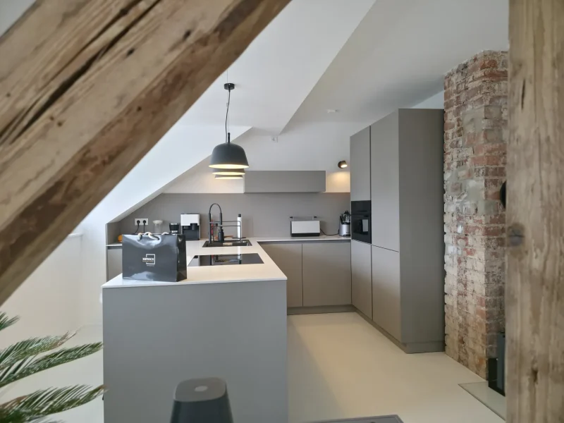 Moderne Küche mit grauen Schränken, einer großen Kücheninsel, einer Säule aus Sichtziegeln, einer schrägen Decke, Hängelampen und Holzbalken. Auf der Kücheninsel stehen Einkaufstaschen. Der Raum wirkt hell und stilvoll – perfekt für alle, die auf Immscout nach ihrem neuen Zuhause suchen oder bereit sind, neue Möglichkeiten des modernen Wohnens zu entdecken.