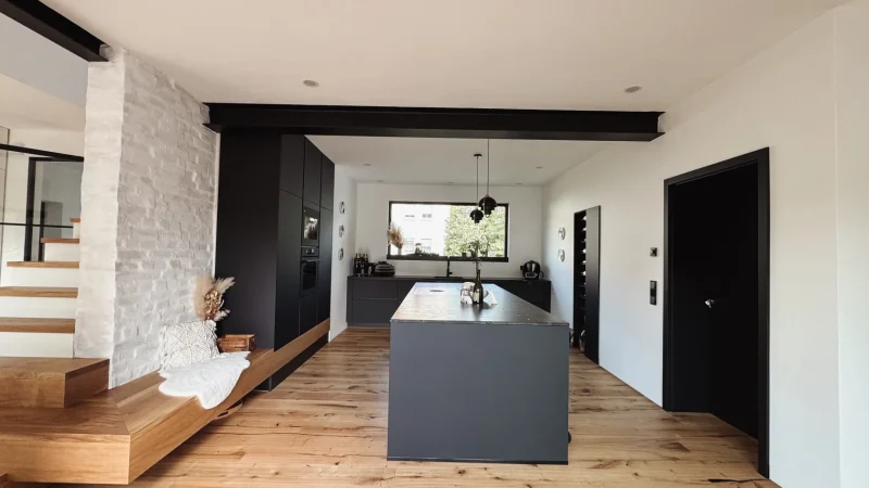 Eine moderne Küche mit schwarzen Schränken, einer großen Kochinsel und Holzboden – perfekt für Ihr neues Zuhause. Links befinden sich eine weiße Ziegelwand und eine Holztreppe. Über der Kochinsel hängen Pendelleuchten, und ein Fenster lässt Tageslicht herein, wodurch neue Möglichkeiten entstehen, jeden Moment zu Hause zu genießen.