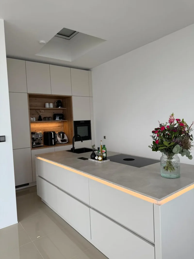Moderne Küche mit weißen Schränken, einer Arbeitsplatte aus Beton, Einbaugeräten und einer Vase mit bunten Blumen auf der Kücheninsel. Im Hintergrund sind Regale mit Küchenutensilien und Flaschen zu sehen. Geplant und realisiert von xanocs Küchen in Leipzig und Grimma