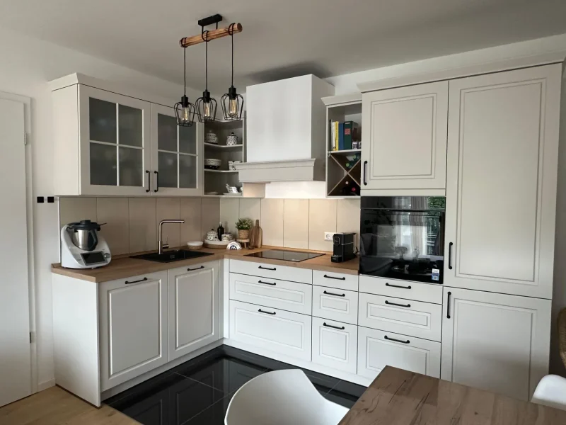 Moderne Küche mit weißen Schränken, Arbeitsplatten aus Holz, Einbaubackofen, Induktionskochfeld, Spüle, Kleingeräten, offenen Regalen und Pendelleuchten darüber. Der Raum wirkt aufgeräumt, übersichtlich und stilvoll – und bietet neue Möglichkeiten für alle, die auf Immscout nach ihrem neuen Zuhause suchen.