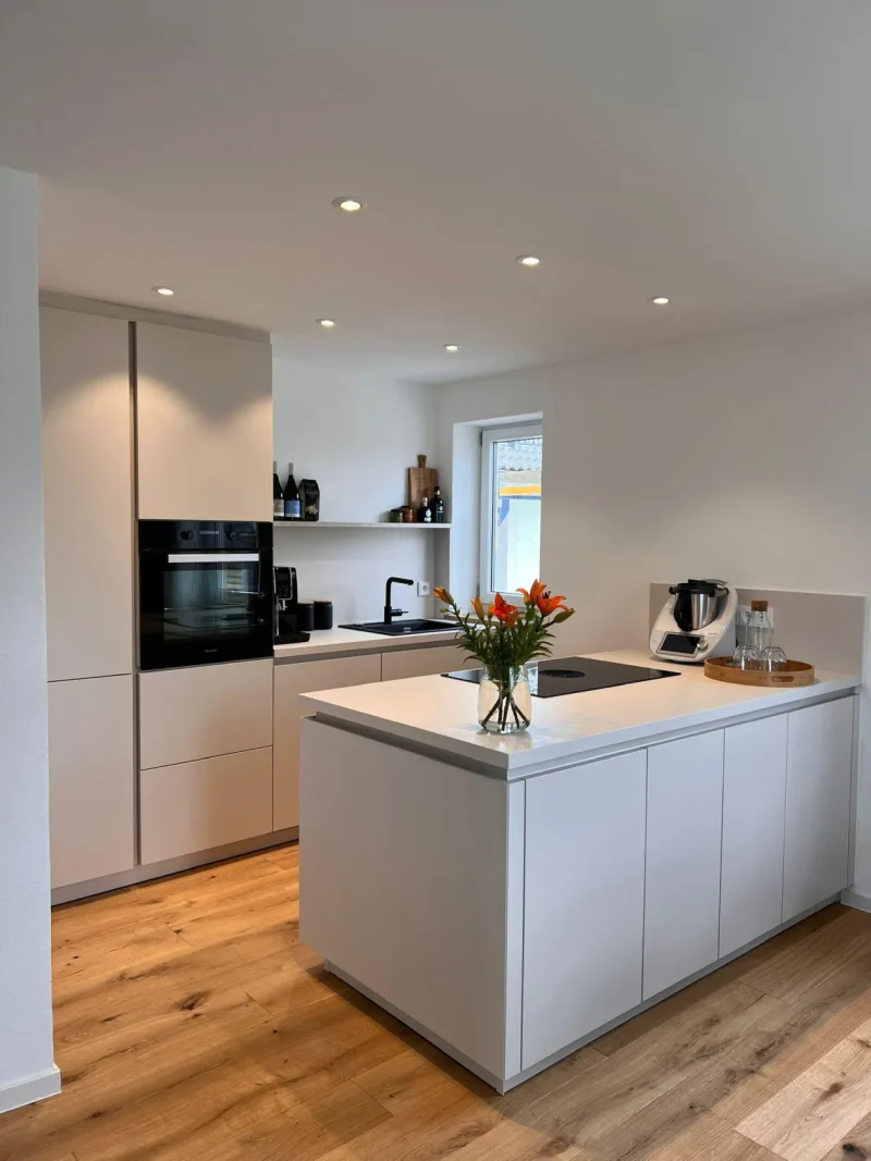 Moderne Küche mit weißen Schränken, Holzboden, Einbaubackofen, Kaffeemaschine und einer weißen Kücheninsel mit einer Vase voller orangefarbener Blumen. Auf den Regalen stehen Gläser und Tassen; durch die Fenster fällt Tageslicht herein – alles, was Sie brauchen, um neue Möglichkeiten in Ihrem neuen Zuhause zu genießen. Entdecken Sie diesen einladenden Raum noch heute auf Immscout.