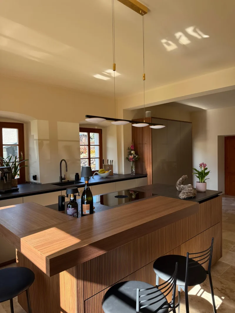 Eine moderne Küche mit einer Kücheninsel aus Holz, schwarzen Barhockern, Pendelleuchten, Pflanzen, Weinflaschen und großen Fenstern, durch die Tageslicht hereinströmt – perfekt, um mit Immscout neue Möglichkeiten zu entdecken und Ihr neues Zuhause zu gestalten.