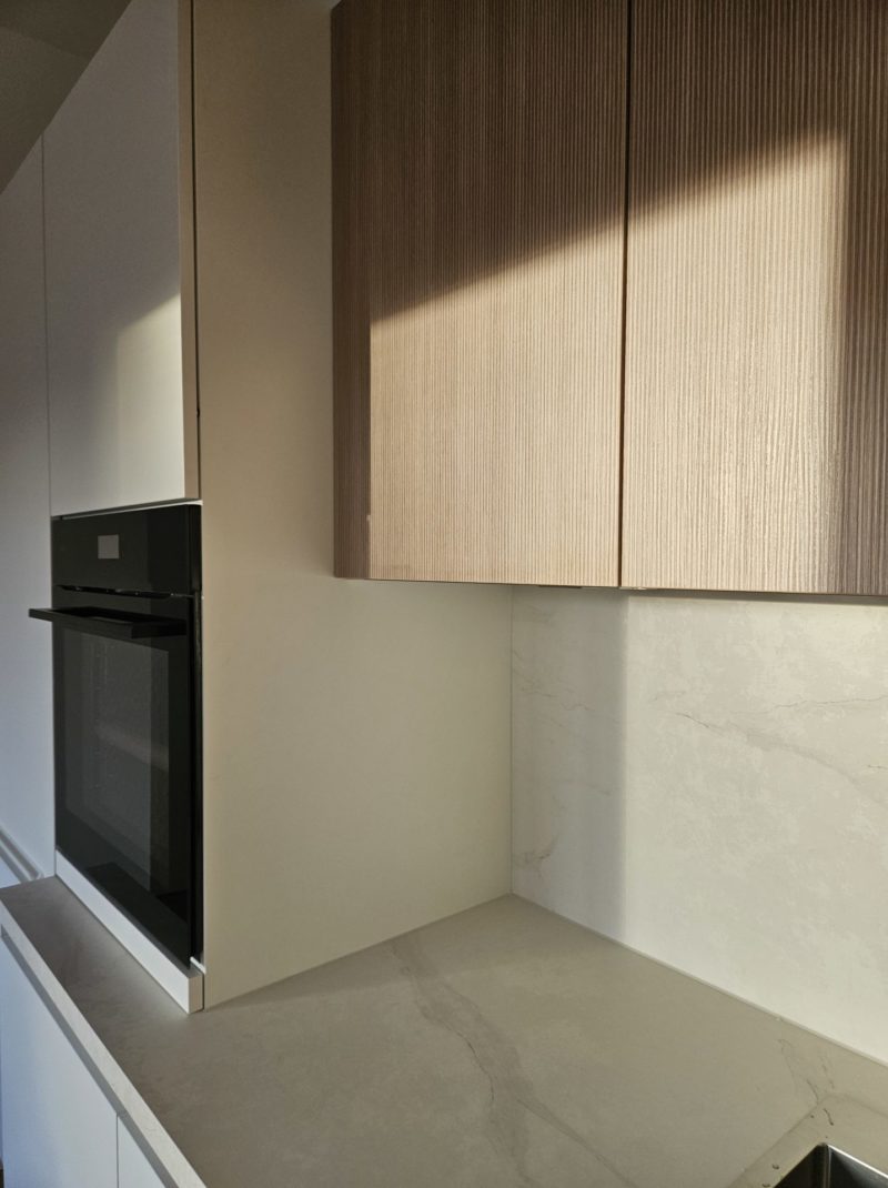 Sonnenlicht fällt auf eine moderne Küchenecke mit hellen Holzschränken, einer weißen Marmor-Rückwand und einem eingebauten schwarzen Backofen. Die klare und aufgeräumte Arbeitsplatte macht diesen Raum zu einer wahren Inspirationsquelle für alle, die von ihrer eigenen Traumküche träumen.
