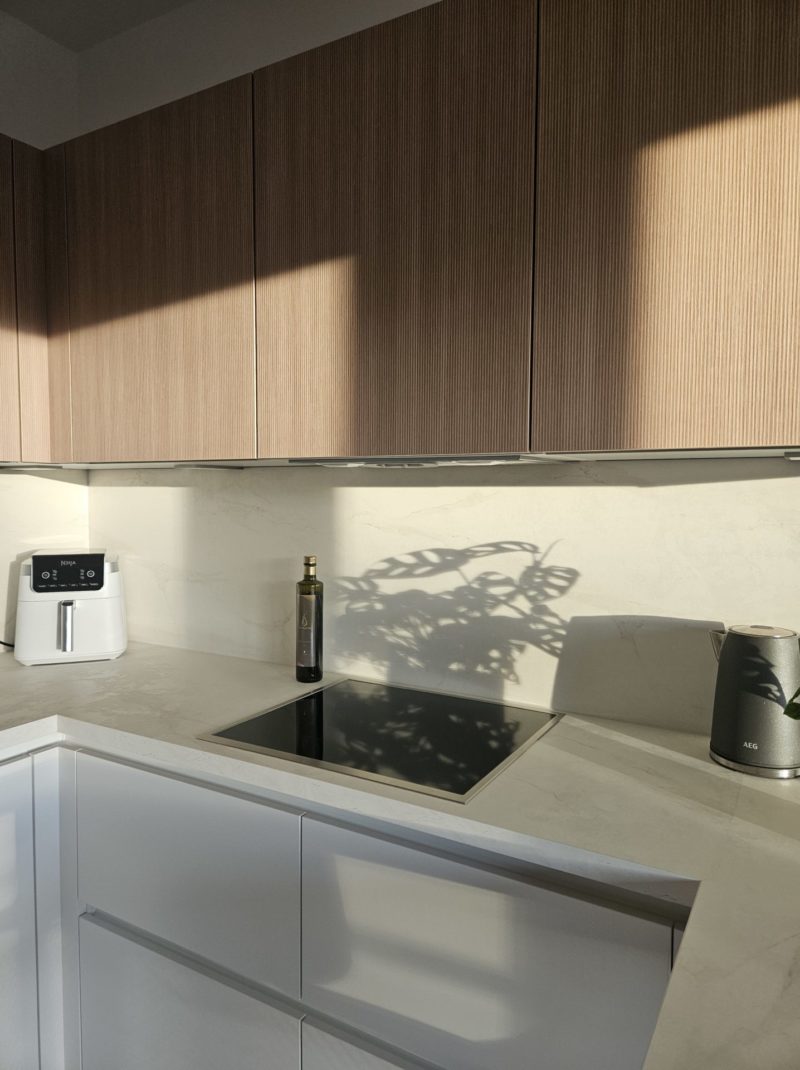 Minimalistische Küche mit hellen Holzschränken, weißer Arbeitsplatte, eingebautem schwarzem Kochfeld, einer Heißluftfritteuse, einer Olivenölflasche und einem Lautsprecher. Das Sonnenlicht wirft den Schatten einer Pflanze auf die Küchenrückwand und macht diesen Raum zu einer wahren Inspiration für Ihre Traumküche.
