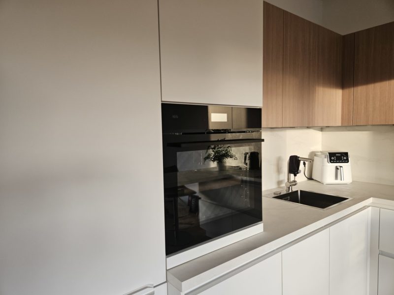 Eine moderne Küche mit schlichten weißen und holzfarbenen Schränken, einem Einbaubackofen, einer Heißluftfritteuse, einer Spüle, einem Schneidebrett und einer Topfpflanze auf der Arbeitsplatte – Ihre tägliche Inspiration für eine echte Traumküche. Sonnenlicht wirft sanfte Schatten auf die Oberflächen und hebt jedes elegante Detail hervor.