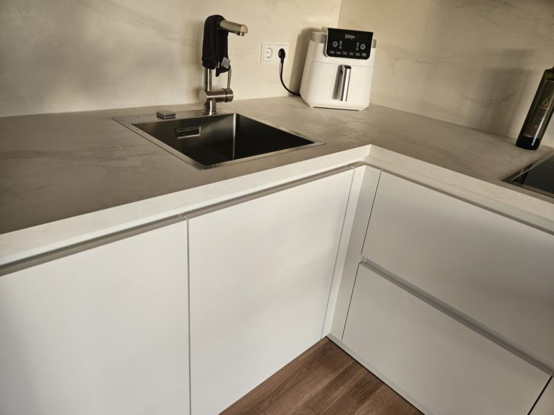 Moderne Küchenecke mit einer Edelstahlspüle, weißen Schränken, einer beigen Arbeitsplatte, einer schwarzen Armatur, einer weißen Heißluftfritteuse und einem Handtuch, das an der Armatur hängt. Diese inspirierende Küche bietet alles für deine Traumküche – am rechten Rand ist teilweise ein dunkles Gerät zu sehen.