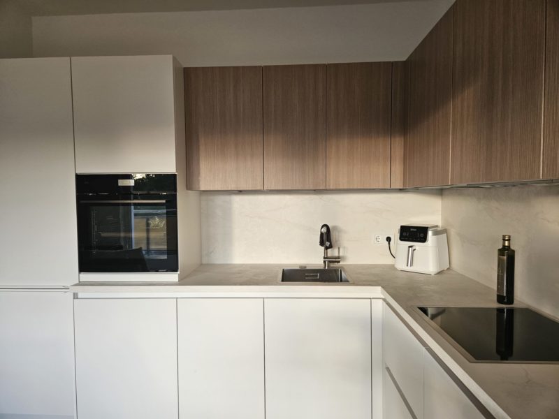 Moderne Küche mit weißen Schränken, Oberschränken aus Holz, einem Einbaubackofen, einer Spüle, einer Heißluftfritteuse, einem Elektrokochfeld und einer Flasche Olivenöl auf der Arbeitsplatte. Der Raum besticht durch klare Linien und minimalistische Inspiration für Ihre Traumküche.
