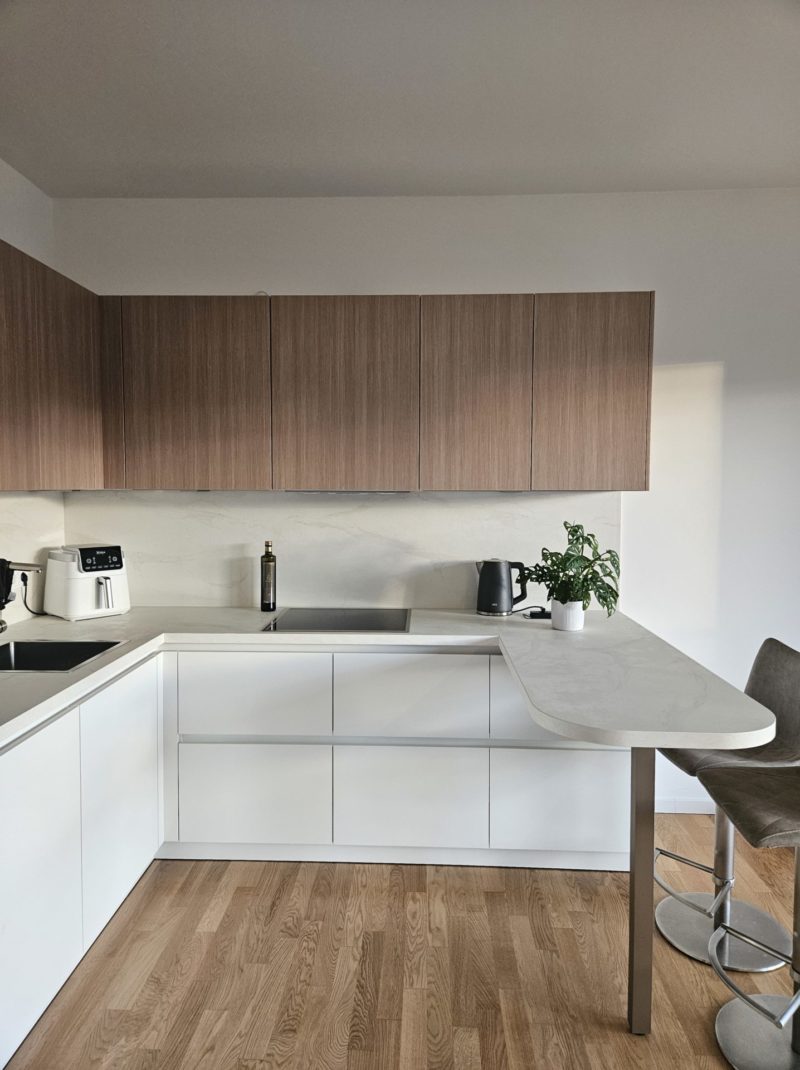 Eine moderne Küche mit weißen Schränken, Oberschränken aus Holz, einer Spüle, Küchengeräten, einer Topfpflanze und einer geschwungenen Arbeitsplatte mit einem Stuhl auf einem Holzboden – ein inspirierender Raum, der Ihre Traumküche zum Leben erweckt.