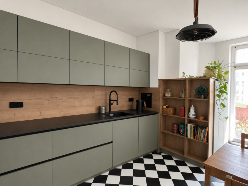 Eine moderne Küche mit grünen Schränken, schwarzer Arbeitsplatte, einer Küchenrückwand aus Holz, einer schwarzen Armatur, offenen Regalen mit Büchern und Pflanzen, einem schwarz-weiß karierten Boden und einem Fenster, das Tageslicht hereinlässt – durchdachte Küchenplanung schafft mehr Raum für Stil und Funktionalität in dieser einladenden Küche.