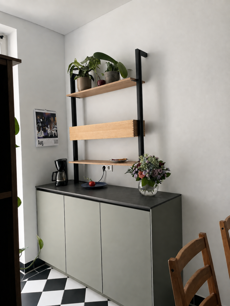 Eine moderne Küchenecke mit einem grauen Schrank, zwei Holzregalen mit Zimmerpflanzen, einer Kaffeemaschine, einer Obstschale, einer Vase mit Blumen und einem Wandkalender, der auf den August aufgeschlagen ist. Durch geschickte Küchenplanung wird der Platz in diesem einladenden Raum optimal genutzt, ergänzt durch schwarz-weiße Fliesenböden.