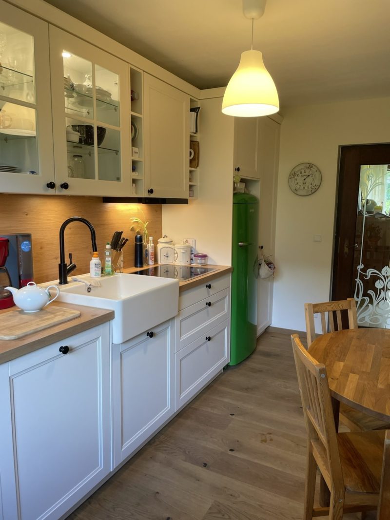 Eine gemütliche Landhausküche mit weißen Schränken, einer Spüle im Landhausstil, einer Arbeitsplatte aus Holz, einem grünen Kühlschrank und einem runden Holztisch mit Stühlen. Auf der Arbeitsplatte stehen Küchenutensilien, an der Wand hängt eine Uhr – jedes Detail trägt dazu bei, ein wahres Stauraumwunder für ein praktisches und zugleich charmantes Wohnen zu schaffen.