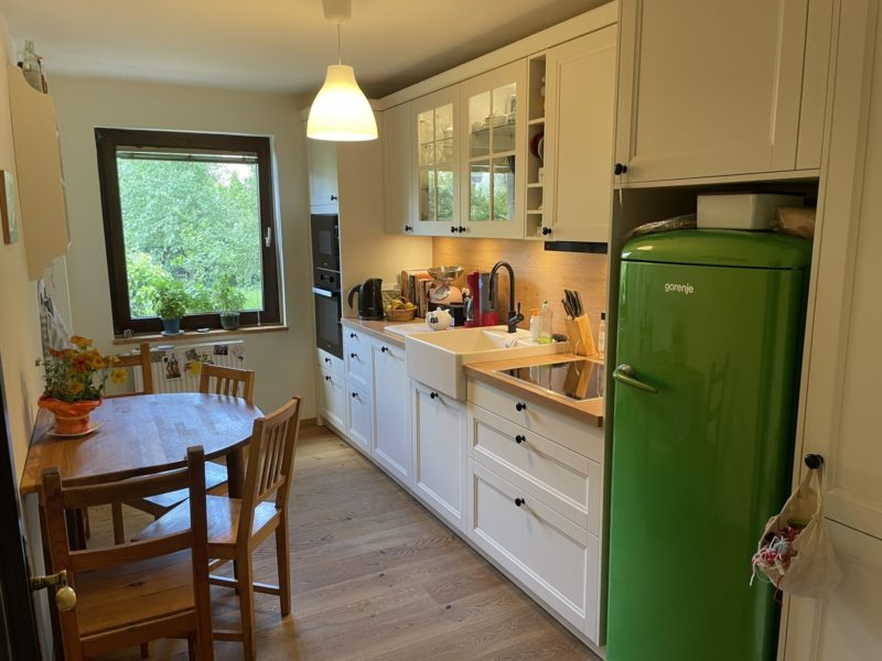 Eine gemütliche Landhausküche mit weißen Schränken, einem grünen Kühlschrank, einem Esstisch aus Holz mit Stühlen, einer Spüle und einem Fenster, durch das man das Grün draußen sehen kann. Auf der Arbeitsplatte und in den Regalen stehen verschiedene Küchenutensilien, was diesen Raum zu einem wahren Stauraumwunder für all Ihre Kochutensilien macht.