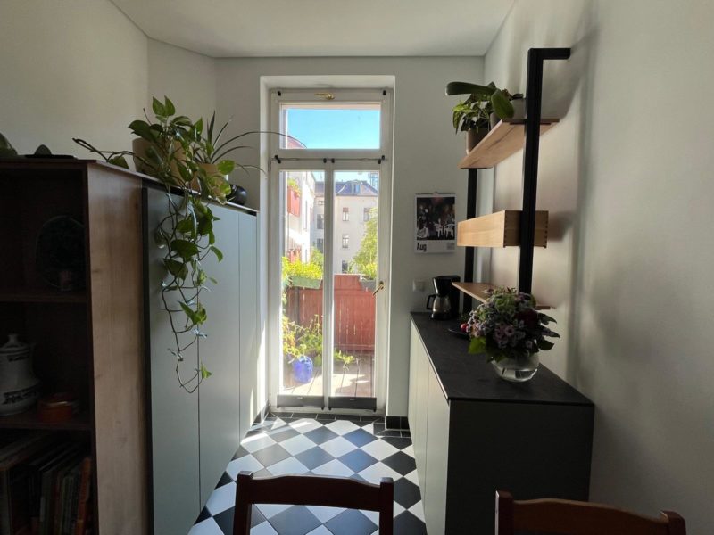 Eine helle Küche mit schwarz-weiß kariertem Boden, Pflanzen auf Regalen und Schränken, einem Blumenstrauß auf der Arbeitsplatte und einer Glastür, die in einen sonnendurchfluteten Garten führt, zeigt, wie eine durchdachte Küchenplanung mehr Raum und Charme in Ihr Zuhause bringen kann.