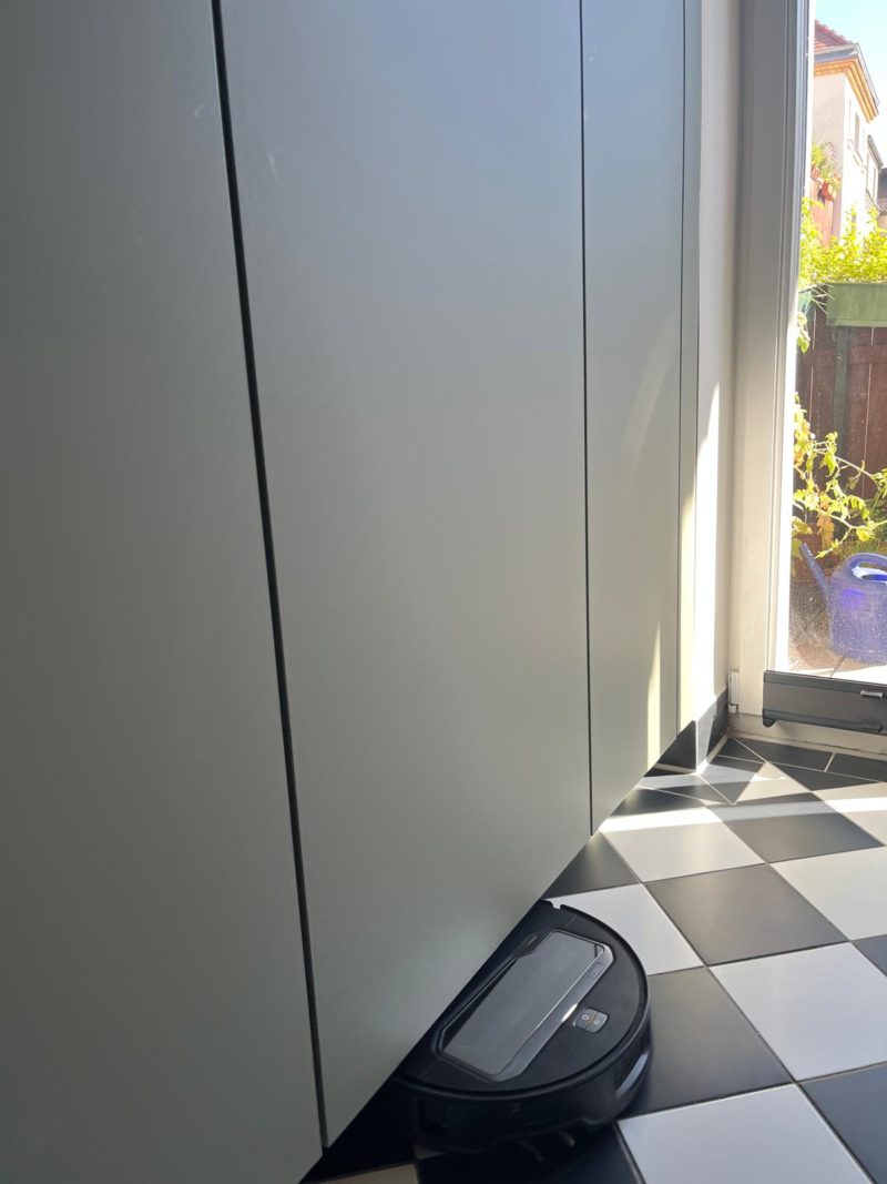 Ein schwarzer Staubsaugerroboter reinigt einen schwarz-weiß karierten Fliesenboden neben weißen Schränken und einer Glastür, durch die Sonnenlicht hereinströmt, und sorgt so durch moderne Küchenplanung für mehr Platz und Effizienz in der Küche.