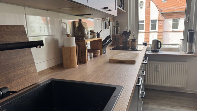 Eine moderne Küche mit Arbeitsplatten aus Holz, die reichlich Arbeitsfläche bieten, einer schwarzen Spüle, verschiedenen Küchenutensilien, Schneidebrettern und Küchengeräten. Sonnenlicht strömt durch ein Fenster herein und erhellt den aufgeräumten und übersichtlichen Raum mit durchdacht gestaltetem Stauraum.