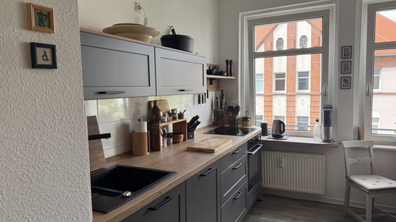 Eine moderne Küche mit grauen Schränken, Arbeitsplatten aus Holz, die reichlich Arbeitsfläche bieten, diversem Küchenzubehör, einem Herd und einer Spüle. Große Fenster lassen Tageslicht herein, und unter dem Fenster stehen ein Stuhl und ein kleiner Heizstrahler. An der Wand hängen Kunstwerke, während durchdachte Stauraumlösungen dafür sorgen, dass alles ordentlich aufgeräumt ist.