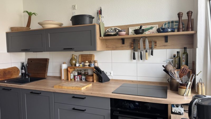 Moderne Küche mit dunklen Schränken, großzügigem Stauraum, einer Arbeitsplatte aus Holz mit reichlich Arbeitsfläche, Regalen für Geschirr und Gewürze, Messern an einer Magnetleiste, Küchenutensilien in Gläsern, Schneidebrettern und einem Kochfeld unter einem Fenster mit Tageslicht.