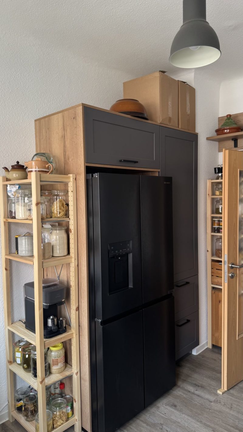 Eine moderne Küchenecke mit einem schwarzen Kühlschrank, reichlich Stauraum auf Holzregalen, auf denen Gläser und Behälter stehen, sowie Kisten und Küchenutensilien, die ordentlich auf den Schränken angeordnet sind. Über der Arbeitsfläche hängt eine Pendelleuchte von der Decke herab.