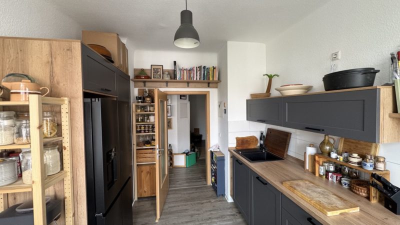 Eine moderne Küche mit schwarzen Schränken, Holzregalen, Vorratsgläsern, einem schwarzen Kühlschrank und einer kleinen Spüle. Über der Tür befinden sich Kochbücher, und die Küchenutensilien sind überall ordentlich verstaut – das bietet clevere Stauraumlösungen und reichlich Arbeitsfläche für eine mühelose Zubereitung von Mahlzeiten.