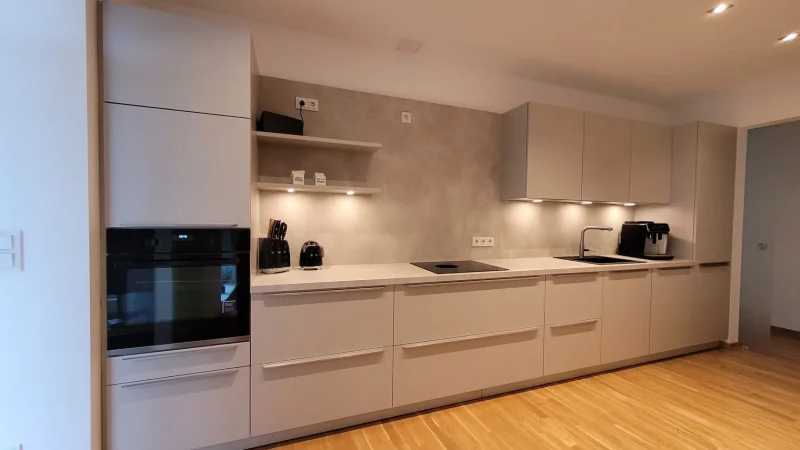 Moderne Küche mit beigen Schränken, schwarzen Geräten, Einbaubackofen, offenen Regalen, hellem Holzboden und Unterbauleuchten entlang einer langen Arbeitsplatte. Der Raum wirkt aufgeräumt und minimalistisch – perfekt für alle, die in ihrem neuen Zuhause nach neuen Möglichkeiten suchen.
