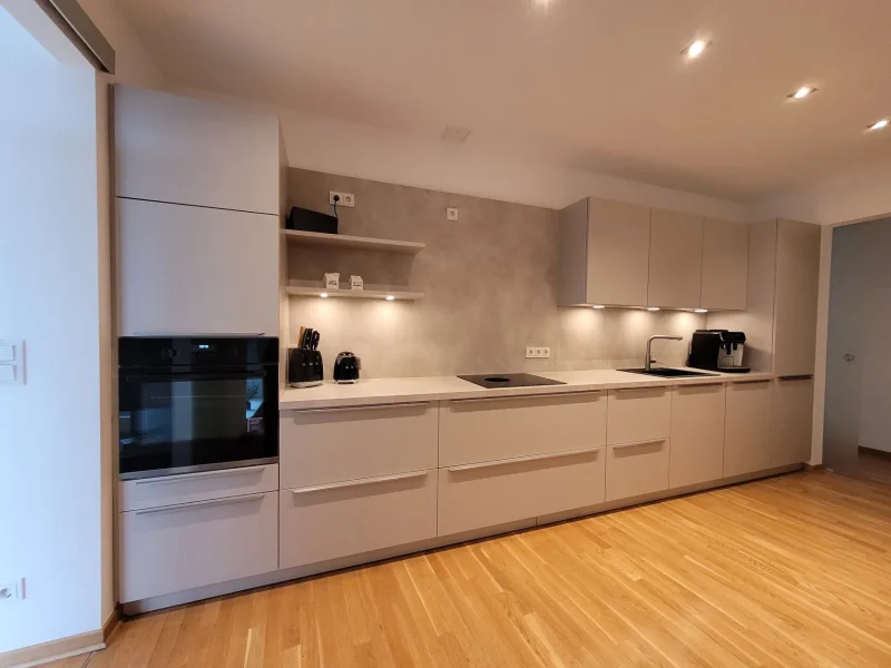 Moderne Küche mit beigen Schränken, schwarzen Geräten, Einbaubackofen, offenen Regalen, hellem Holzboden und Unterbauleuchten entlang einer langen Arbeitsplatte. Der Raum wirkt aufgeräumt und minimalistisch – perfekt für alle, die in ihrem neuen Zuhause nach neuen Möglichkeiten suchen.