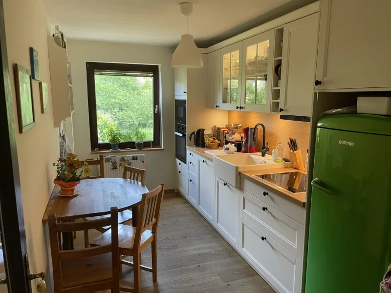 Eine gemütliche Küche mit weißen Schränken, einem grünen Kühlschrank, einem Esstisch aus Holz mit Stühlen und einem Fenster, durch das Tageslicht hereinströmt. Pflanzen und Küchenutensilien schmücken den Raum und schaffen die perfekte Atmosphäre für Ihr neues Zuhause und eröffnen neue Möglichkeiten für ein komfortables Leben.