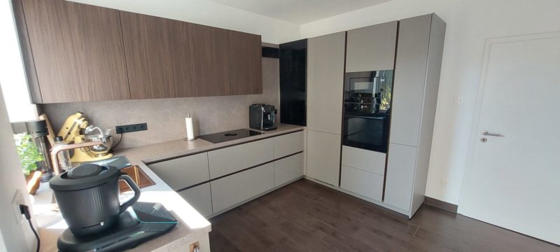 Eine moderne Wohlfühlküche mit Oberschränken in Holztönen, hellgrauen Unterschränken, einem Einbaubackofen und einer Arbeitsfläche mit Einbaugeräten – für einen gelungenen Küchenneustart.