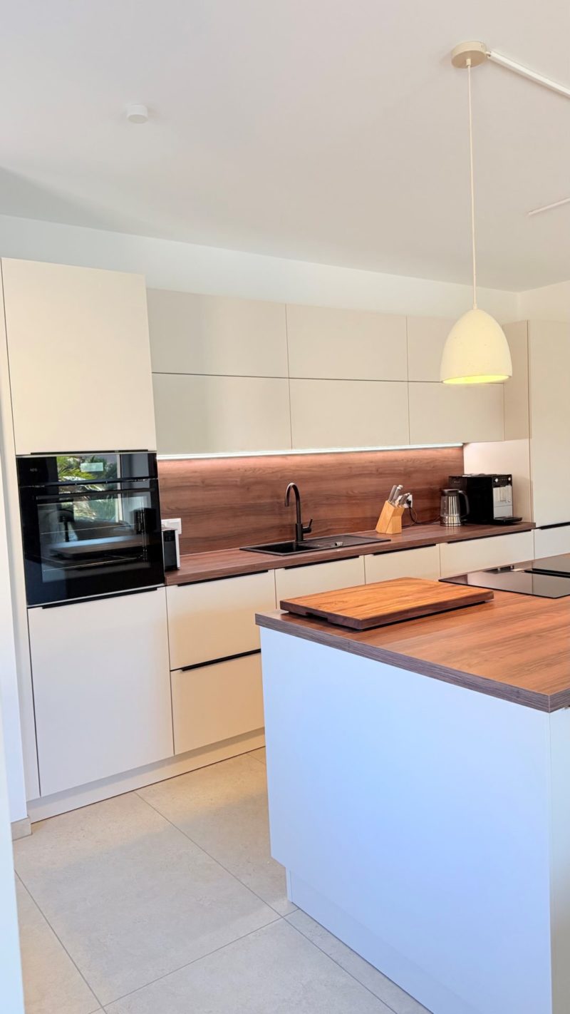 Moderne Küche in einem offenen Wohnbereich mit weißen Schränken, Arbeitsplatten und Fliesenspiegel in Holztönen, einer schwarzen Spüle und einer Kücheninsel in der Mitte.