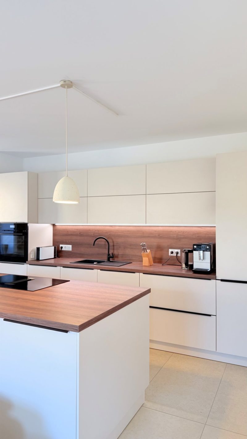 Moderne Küche mit weißen Schränken, einer Arbeitsplatte und einer Rückwand in Holzoptik, einer schwarzen Spüle und einer Kücheninsel in einem offenen Wohnbereich.